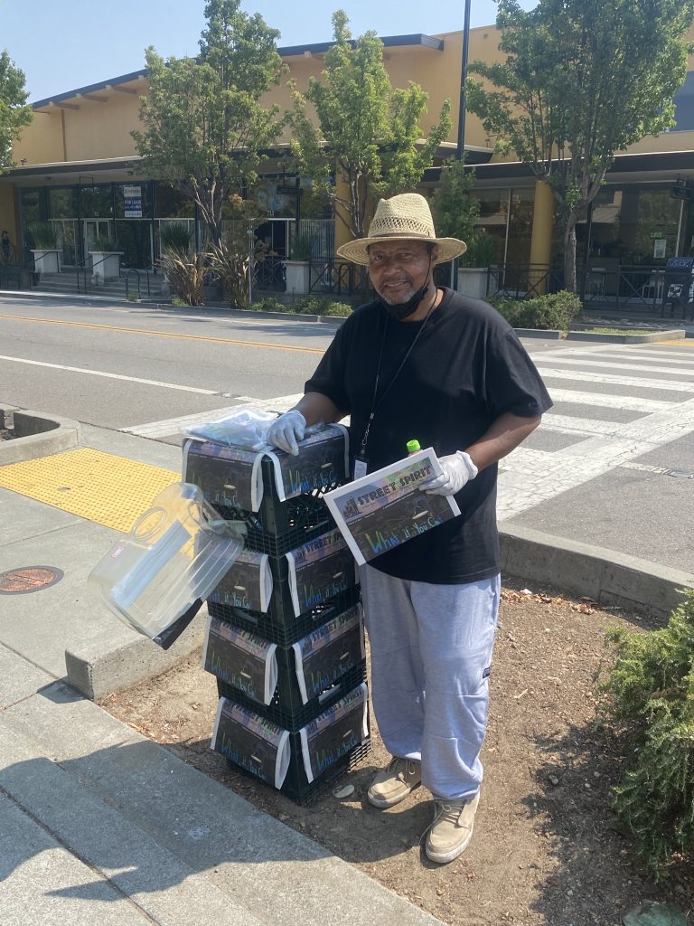 Vernon Dailey stands on the corner with crates displaying his newspaper. He is wearing a black t-shirt, a straw hat, grey sweatpants, and lace up leather sneakers.